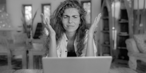 Vrouw kijkt naar een laptop en is duidelijk gefrustreerd en boos. Ze heft haar handen omhoog om de frustratie te benadrukken.