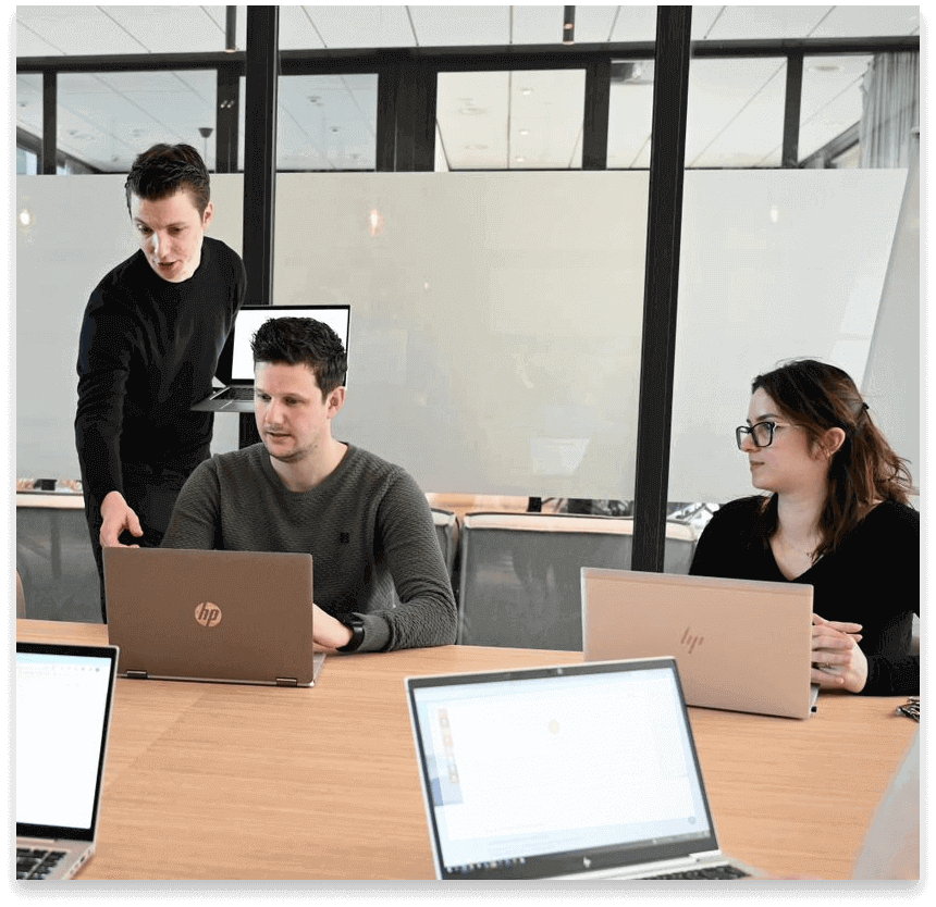 Een man en een vrouw krijgen van een trainer uitleg terwijl ze op hun eigen laptop aan het werk zijn. Op de voorgrond zijn nog twee opengeklapte laptops te zien.